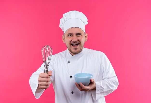 Joyful young handsome cook in chef uniform holding whisk and bowl isolated on pink wall