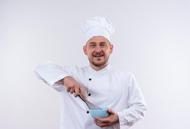 Joyful young handsome cook in chef uniform holding whisk and bowl on isolated white wall