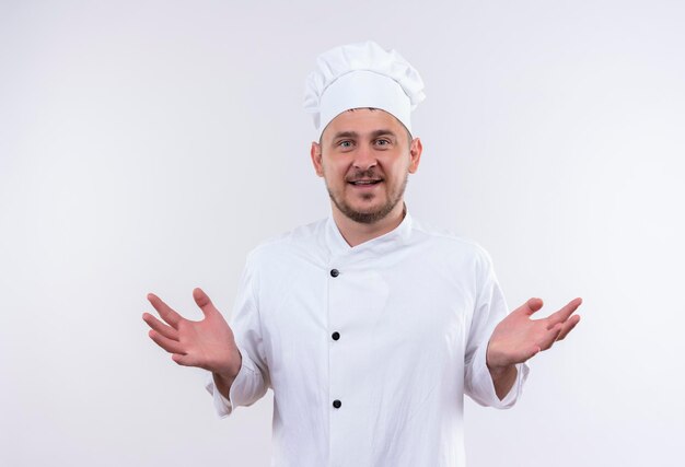 Радостный молодой красивый повар в униформе шеф-повара показывает пустые руки, изолированные на белой стене