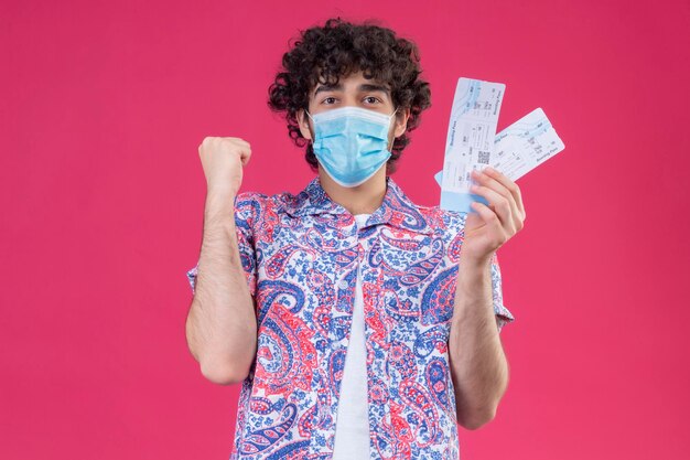 Joyful young handsome curly traveler man wearing medical mask holding airplane tickets with raised fist on isolated pink space with copy space