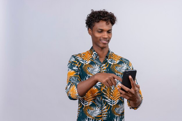 Joyful young handsome dark-skinned man with curly hair in leaves printed shirt holding smartphone hands and touching screen of mobile phone on a white background