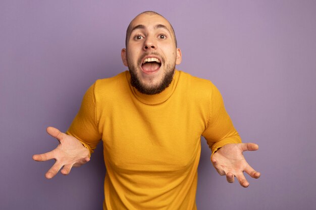 Joyful  young handsome guy spreading hands isolated on purple wall