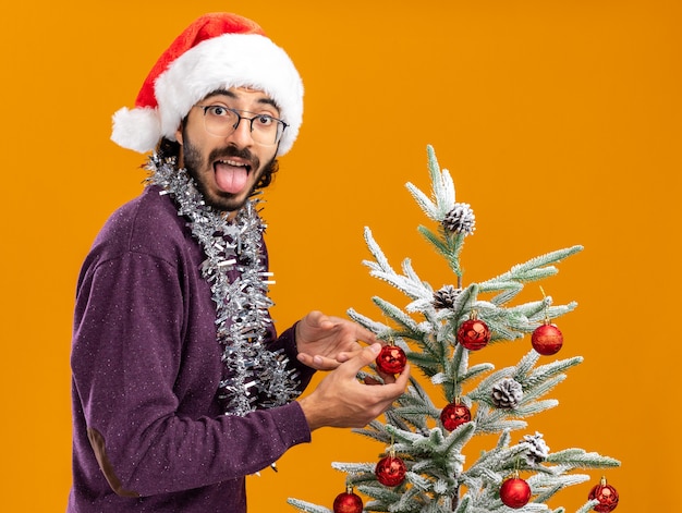 Joyful young handsome guy standing nearby christmas tree wearing christmas hat with garland on neck showing tongue isolated on orange background