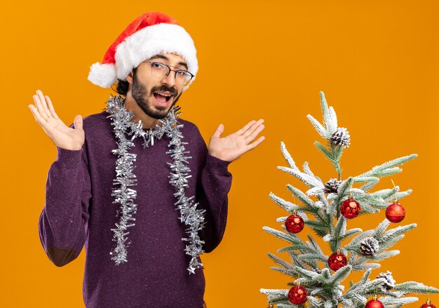 Joyful young handsome guy standing nearby christmas tree wearing christmas hat with garland on neck spreading hands isolated on orange wall