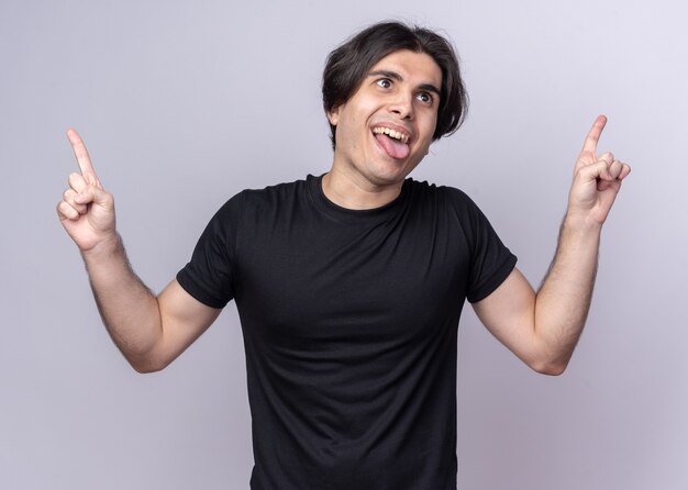 Joyful young handsome guy wearing black t-shirt points at up showing tongue isolated on white wall