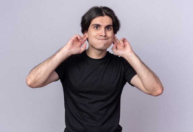 Joyful young handsome guy wearing black t-shirt putting hands on ears isolated on white wall