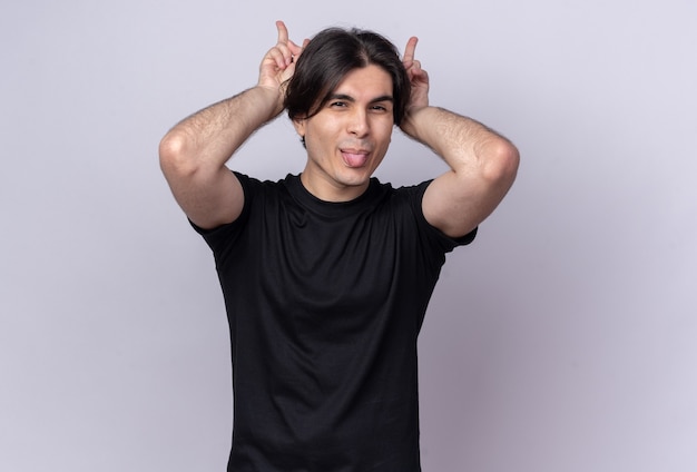 Joyful young handsome guy wearing black t-shirt showing tongue doing bunny ears gesture isolated on white wall