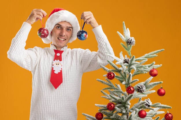 Joyful young handsome guy wearing christmas hat and santa claus tie standing near decorated christmas tree holding christmas ball ornaments near head  isolated on orange wall