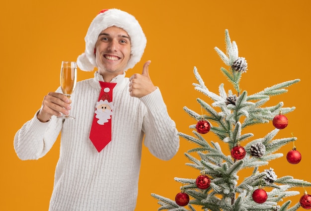 クリスマスの帽子とサンタクロースのネクタイを身に着けているうれしそうな若いハンサムな男は、オレンジ色の壁に分離された親指を示すシャンパンのガラスを保持している装飾されたクリスマスツリーの近くに立っています