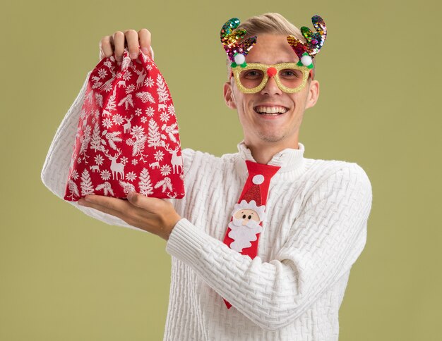クリスマスノベルティメガネとオリーブグリーンの壁に分離されたクリスマス袋を保持しているサンタクロースのネクタイを身に着けているうれしそうな若いハンサムな男