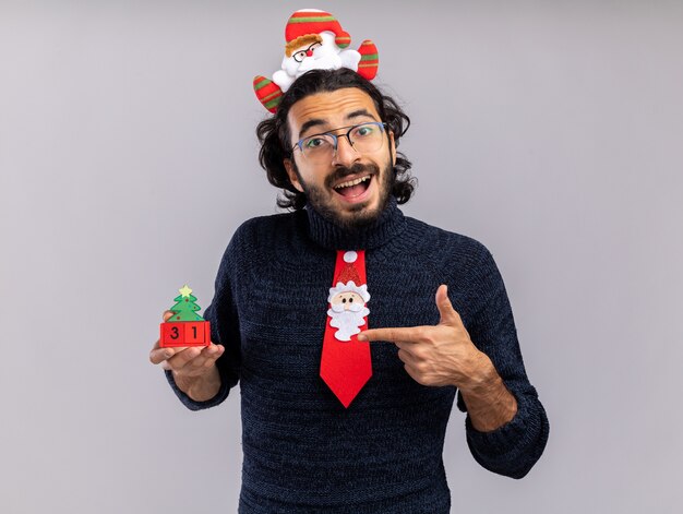 Радостный молодой красивый парень в рождественском галстуке с обручем для волос держит и указывает на рождественскую игрушку, изолированную на белой стене