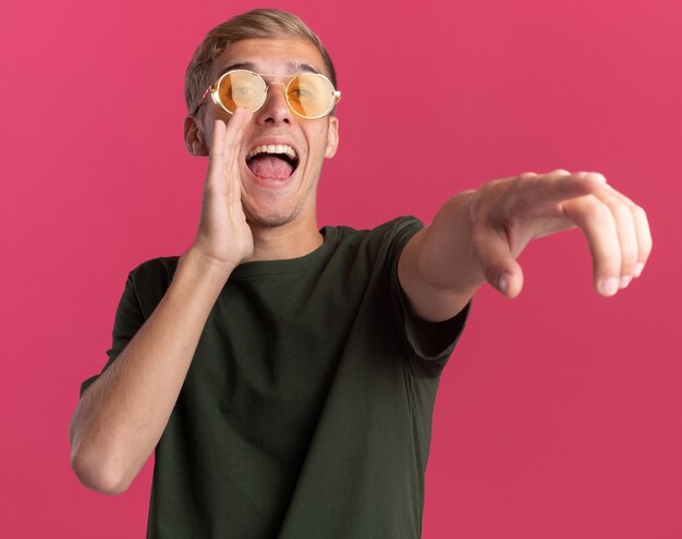 Joyful young handsome guy wearing green shirt and glasses points at side and calling someone isolated on pink wall