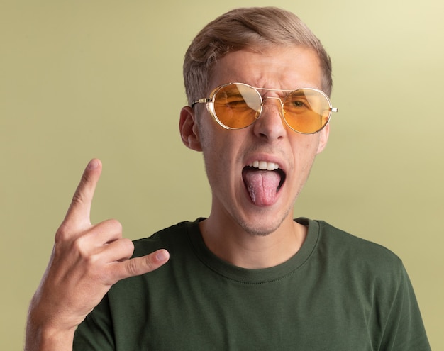 Joyful young handsome guy wearing green shirt with glasses showing goat gesture isolated on olive green wall