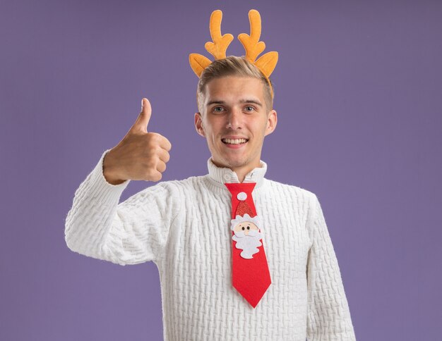 Joyful young handsome guy wearing reindeer antlers headband and santa claus tie  showing thumb up isolated on purple wall