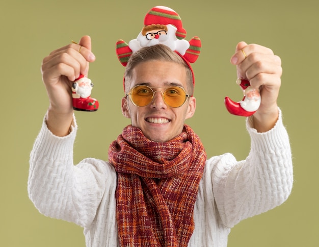 Бесплатное фото Радостный молодой красивый парень в повязке на голову санта-клауса и шарфе, протягивающий рождественские украшения санта-клауса к изолированной на оливково-зеленой стене
