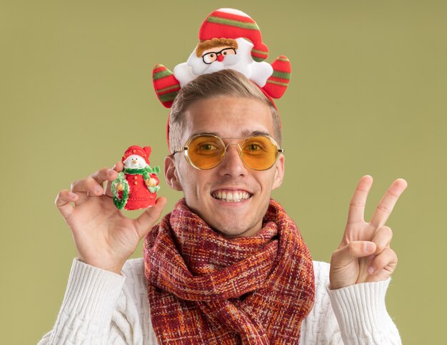 Joyful young handsome guy wearing santa claus headband and scarf  holding snowman christmas ornament doing peace sign isolated on olive green wall