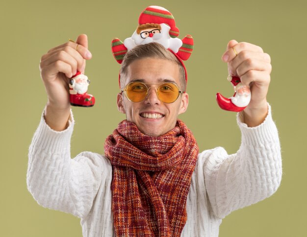 Free photo joyful young handsome guy wearing santa claus headband and scarf  stretching out santa claus christmas ornaments towards isolated on olive green wall