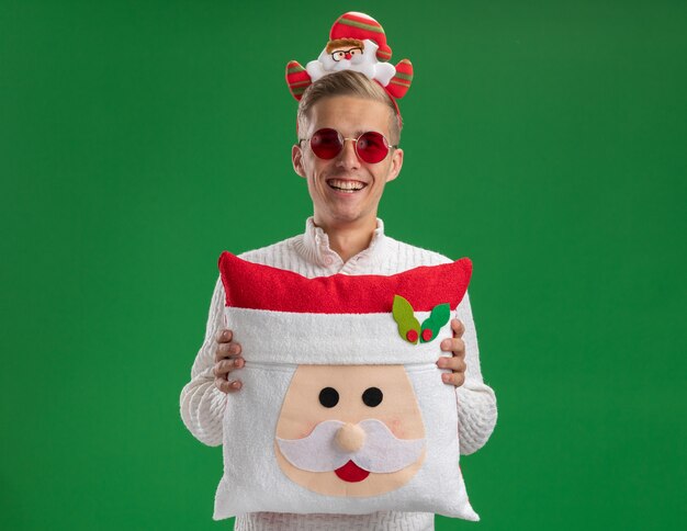 Joyful young handsome guy wearing santa claus headband with glasses holding santa claus pillow  laughing isolated on green wall with copy space
