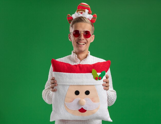 Joyful young handsome guy wearing santa claus headband with glasses holding santa claus pillow looking at camera laughing isolated on green background