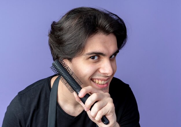 Joyful young handsome male barber in uniform holding comb isolated on blue wall