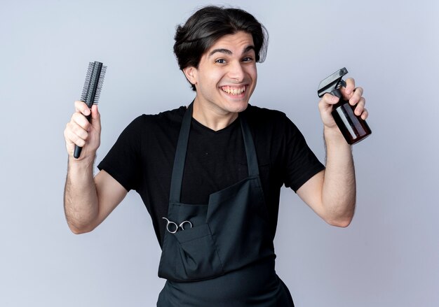 Joyful young handsome male barber in uniform holding comb with spray bottle isolated on white background