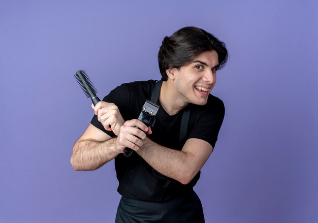 Joyful young handsome male barber in uniform holding and crossing comb with hair clippers isolated on blue background