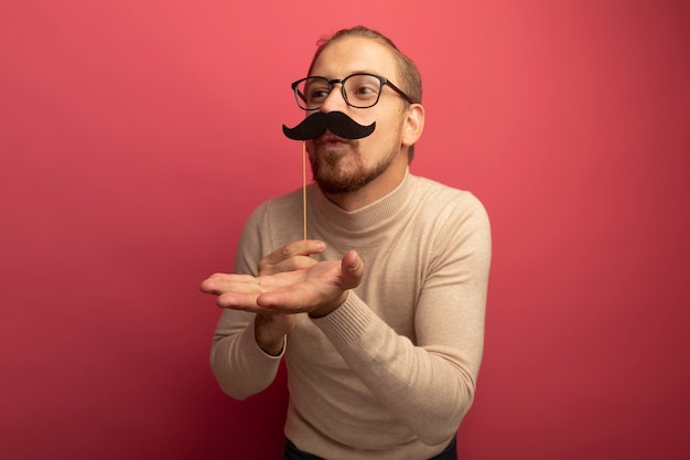 Joyful young handsome man in beige turtleneck and glasses holding funny mustache on stick blowing a kiss 