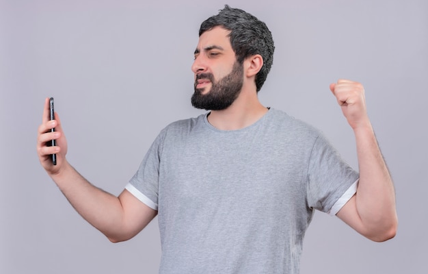 Joyful young handsome man holding and looking at mobile phone and raising fist isolated on white