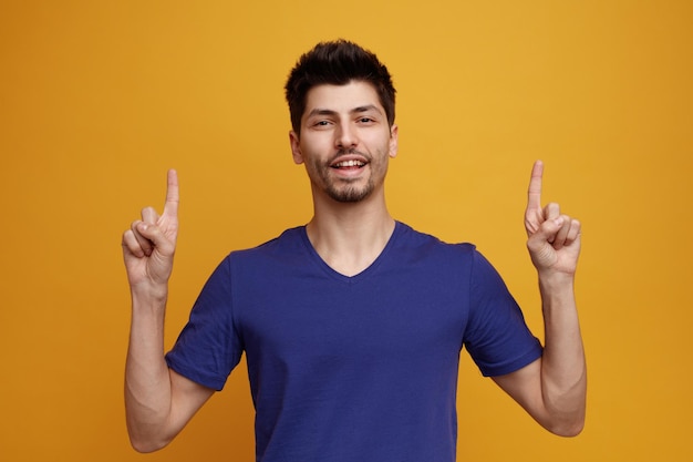 Joyful young handsome man looking at camera pointing up on yellow background