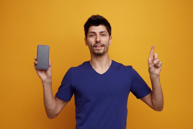 Joyful young handsome man looking at camera showing mobile phone to camera pointing up on yellow background