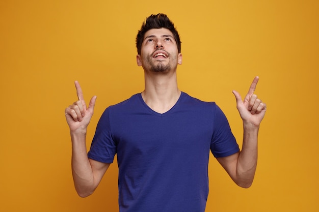 Joyful young handsome man looking and pointing up on yellow background