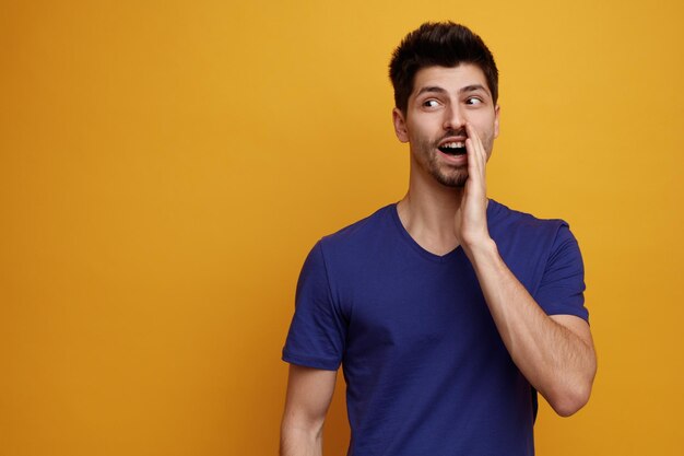 Joyful young handsome man looking at side doing whisper gesture on yellow background with copy space