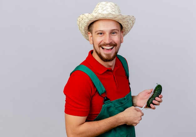 Joyful young handsome slavic gardener in uniform and hat looking holding cucumber and tape meter measuring cucumber isolated