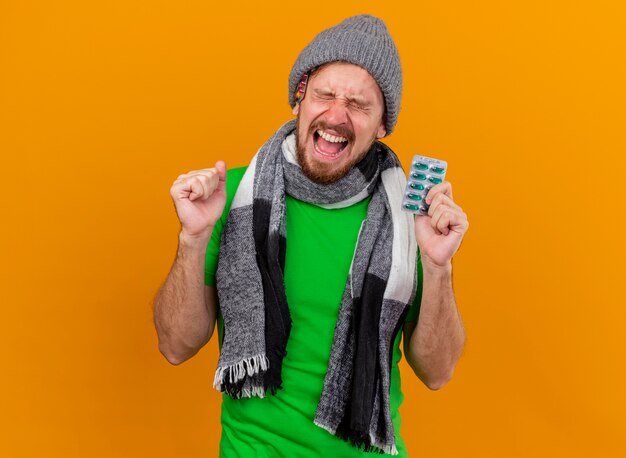 Joyful young handsome slavic ill man wearing winter hat and scarf, holding pack of capsules, doing yes gesture with closed eyes