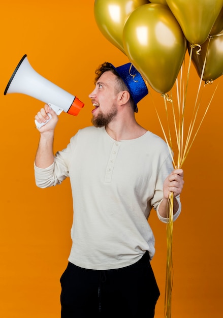 Free photo joyful young handsome slavic party guy wearing party hat holding balloons turning head to side talking by speaker isolated on orange background