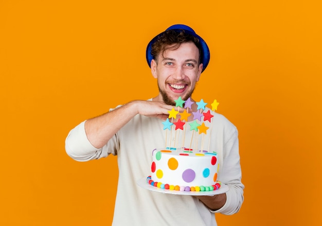 Joyful young handsome slavic party guy wearing party hat holding birthday cake with stars looking at camera keeping hand under chin isolated on orange background with copy space