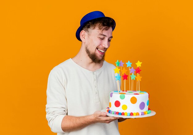 Joyful young handsome slavic party guy wearing party hat holding and looking at birthday cake with stars isolated on orange background with copy space