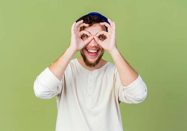 Joyful young handsome slavic party guy wearing party hat looking at camera doing look gesture using hands as binoculars isolated on olive green background with copy space