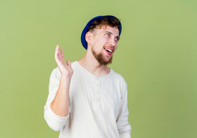 Радостный молодой красивый славянский тусовщик в партийной шляпе смотрит прямо и машет кому-то изолированному на оливково-зеленом фоне с копией пространства