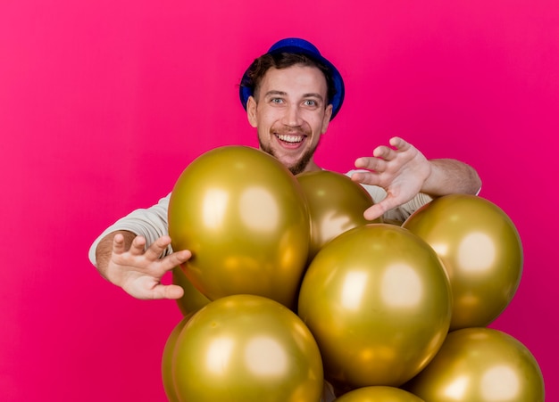 Joyful young handsome slavic party guy wearing party hat standing behind balloons looking at camera showing empty hands isolated on crimson background