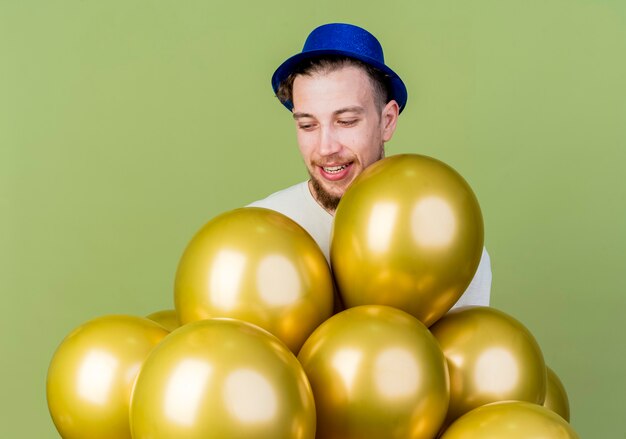 Joyful young handsome slavic party guy wearing party hat standing behind balloons and looking at them isolated on olive green background