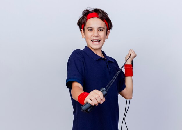 Joyful young handsome sporty boy wearing headband and wristbands with dental braces looking at camera pulling jump rope isolated on white background with copy space