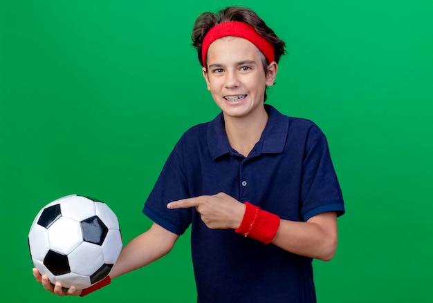 Joyful young handsome sporty boy wearing headband and wristbands with dental braces looking at front holding and pointing at soccer ball isolated on green wall with copy space