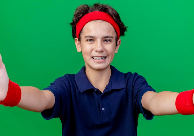 Радостный молодой красивый спортивный мальчик, носящий повязку на голову и браслеты с зубными скобами, смотрит вперед и протягивает руки вперед, изолированные на зеленой стене