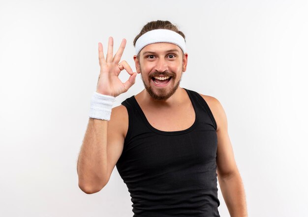 Joyful young handsome sporty man wearing headband and wristbands doing ok sign isolated on white wall