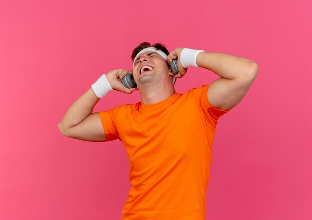 Joyful young handsome sporty man wearing headband and wristbands and headphones holding headphones with closed eyes isolated on pink