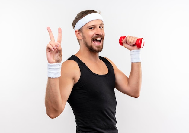 Joyful young handsome sporty man wearing headband and wristbands holding dumbbell and doing peace sign isolated on white wall