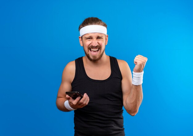 Joyful young handsome sporty man wearing headband and wristbands holding mobile phone and clenching fist isolated on blue wall with copy space