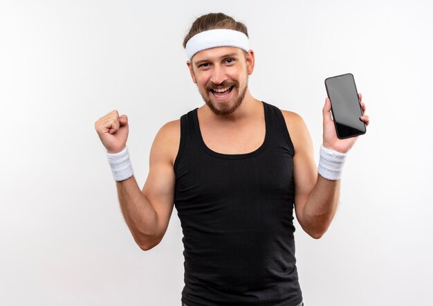 Joyful young handsome sporty man wearing headband and wristbands holding mobile phone and clenching fist isolated on white wall
