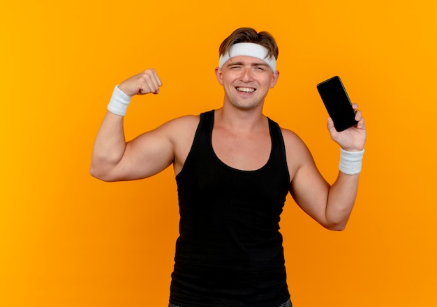 Joyful young handsome sporty man wearing headband and wristbands holding mobile phone and gesturing strong isolated on orange
